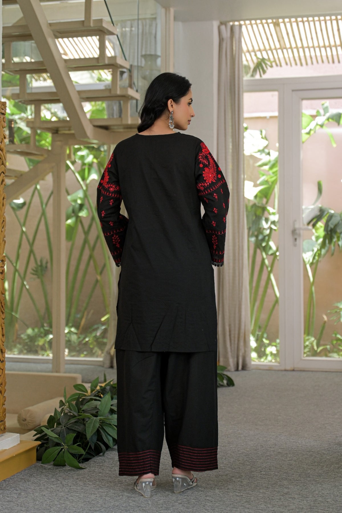 A woman stands with her back to the camera wearing a black three-piece kurta set with red embroidered sleeves and striped trouser hems, in a modern indoor setting with a staircase and glass doors.