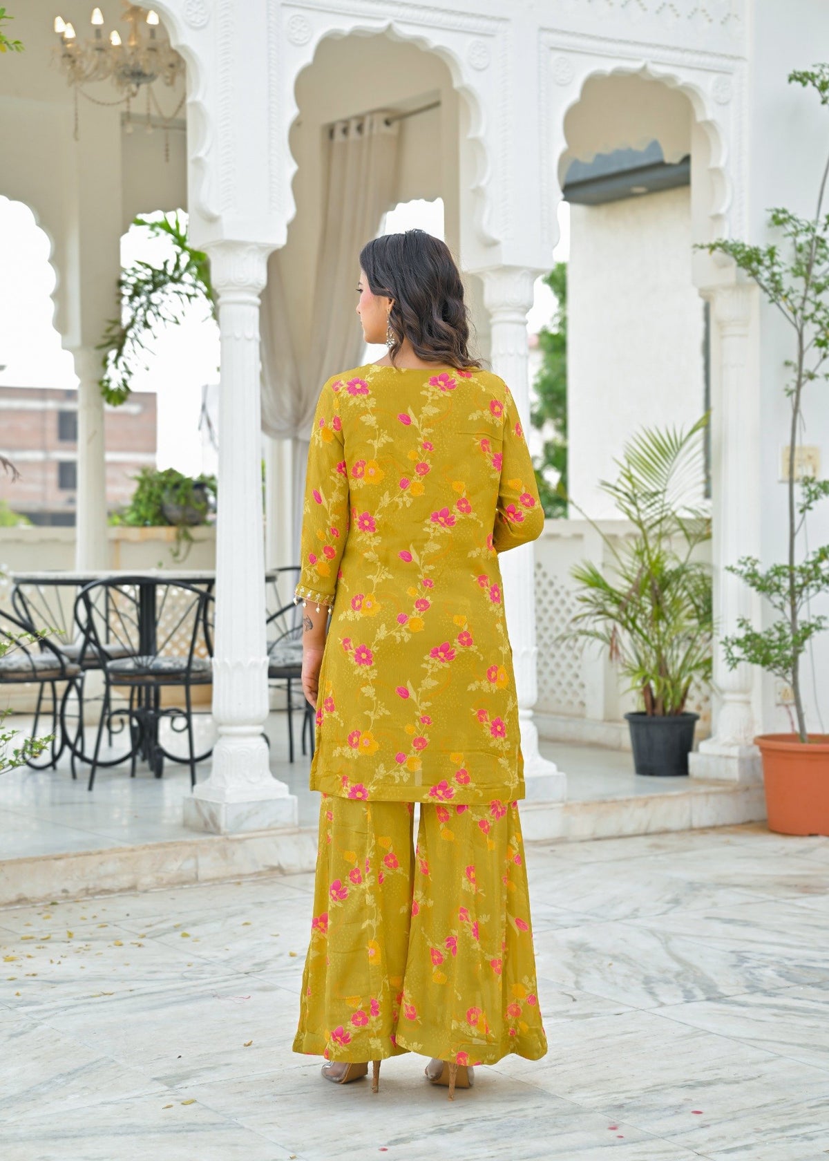 A woman viewed from behind wearing a yellow kurta and wide-leg pants set with a pink and orange floral print, standing in a white arched courtyard.