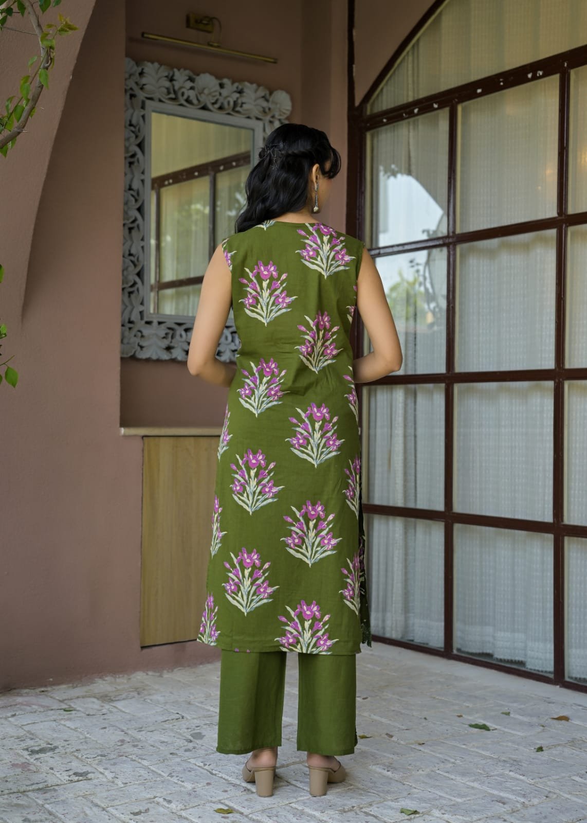 A woman viewed from behind wearing an olive green kurta with pink and white floral prints, paired with matching olive green pants, standing in front of a building with a tiled floor and large window panes.