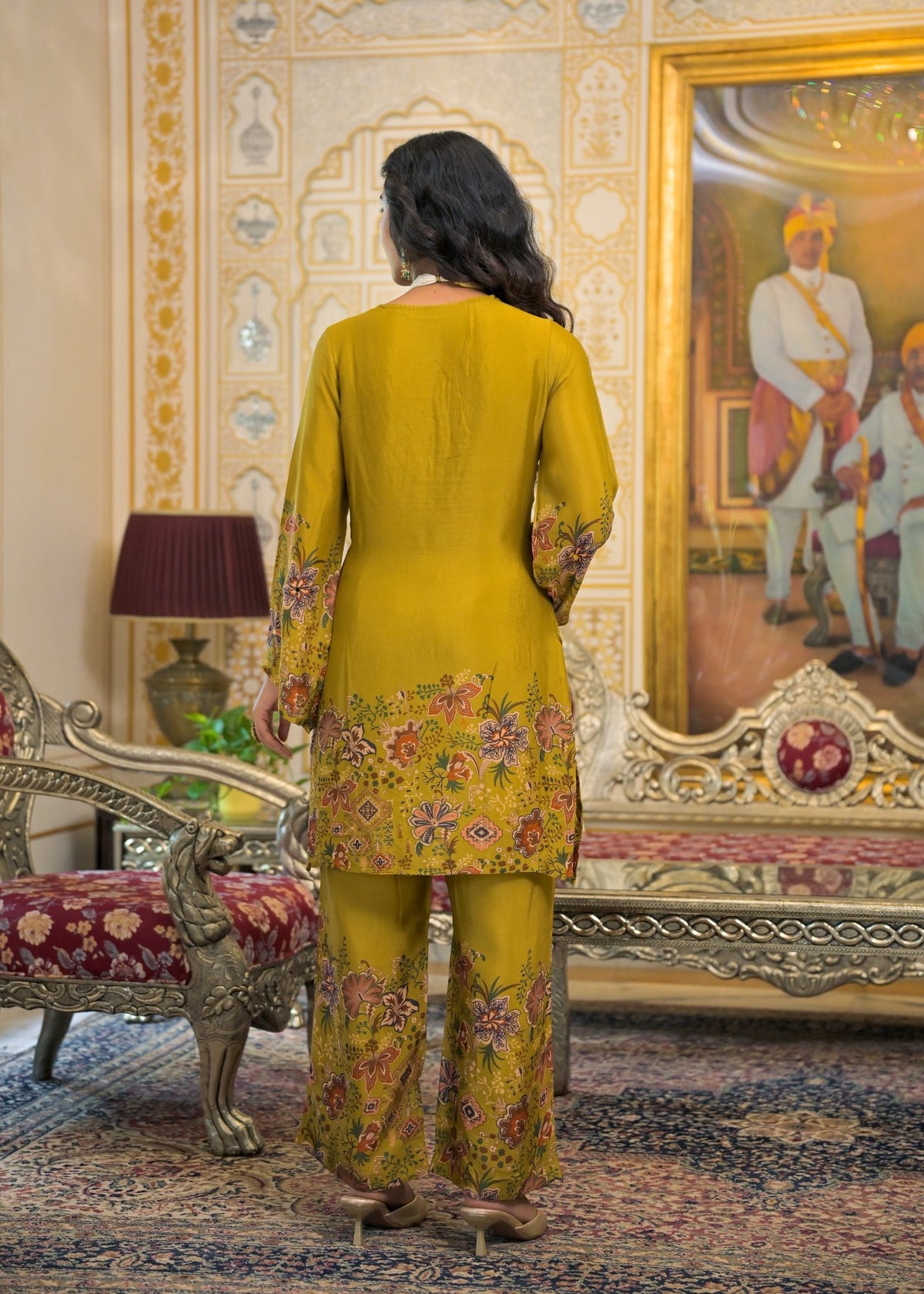 A woman stands with her back to the camera wearing a mustard yellow three-piece traditional outfit with floral embroidery, set in an ornate, vintage-style room with a large painting and patterned furniture.
