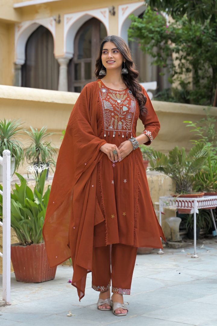 A woman models a rust-orange salwar kameez suit with a matching dupatta in an outdoor courtyard setting. The kurta features white and gold embroidery on the chest, paired with matching pants and a draped dupatta.