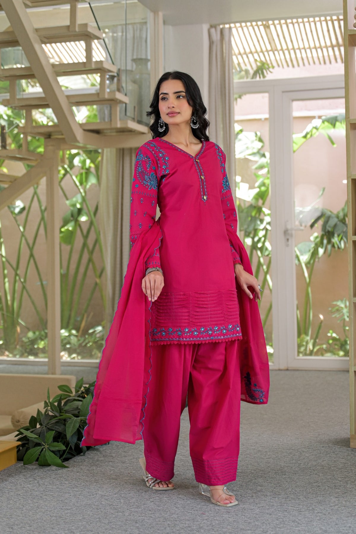 A woman modeling a bright pink traditional three-piece salwar kameez suit with blue embroidery, standing in a modern indoor setting with large windows and plants.