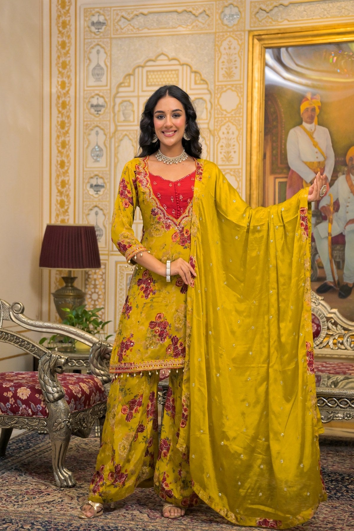 A woman models a traditional Indian salwar kameez suit set in a grand, ornate room. The set features a bright yellow base with pink floral patterns, a red embroidered kameez bodice, and a matching yellow dupatta with subtle gold embroidery.