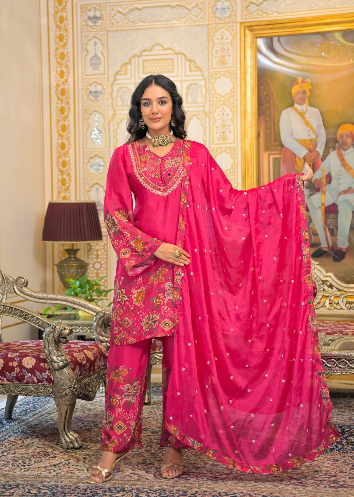 A woman models a bright pink traditional Indian salwar kameez suit with intricate gold and multicolor embroidery, paired with a matching sheer pink dupatta. She is standing in an ornate, traditional Indian room with a large painting on the wall.
