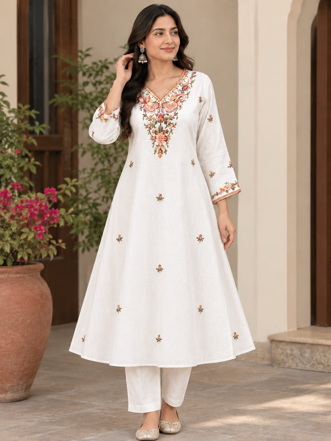 A woman models a white three-piece Indian kurta set with colorful floral embroidery on the neckline and scattered motifs on the front, paired with matching white trousers and beige flat shoes, set against a warm-toned architectural background with a potted plant.