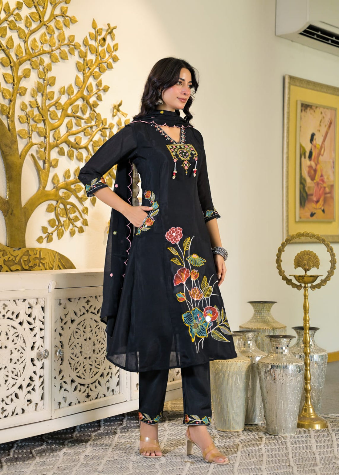 A woman models a black three-piece salwar kameez suit with colorful floral embroidery on the kameez and pants, standing in an indoor setting with decorative gold and white furniture.