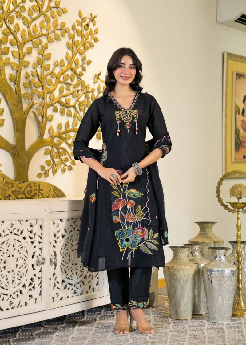 A woman models a black three-piece salwar kameez suit with colorful floral embroidery on the kameez and pants, standing in an indoor setting with decorative gold and white furniture.