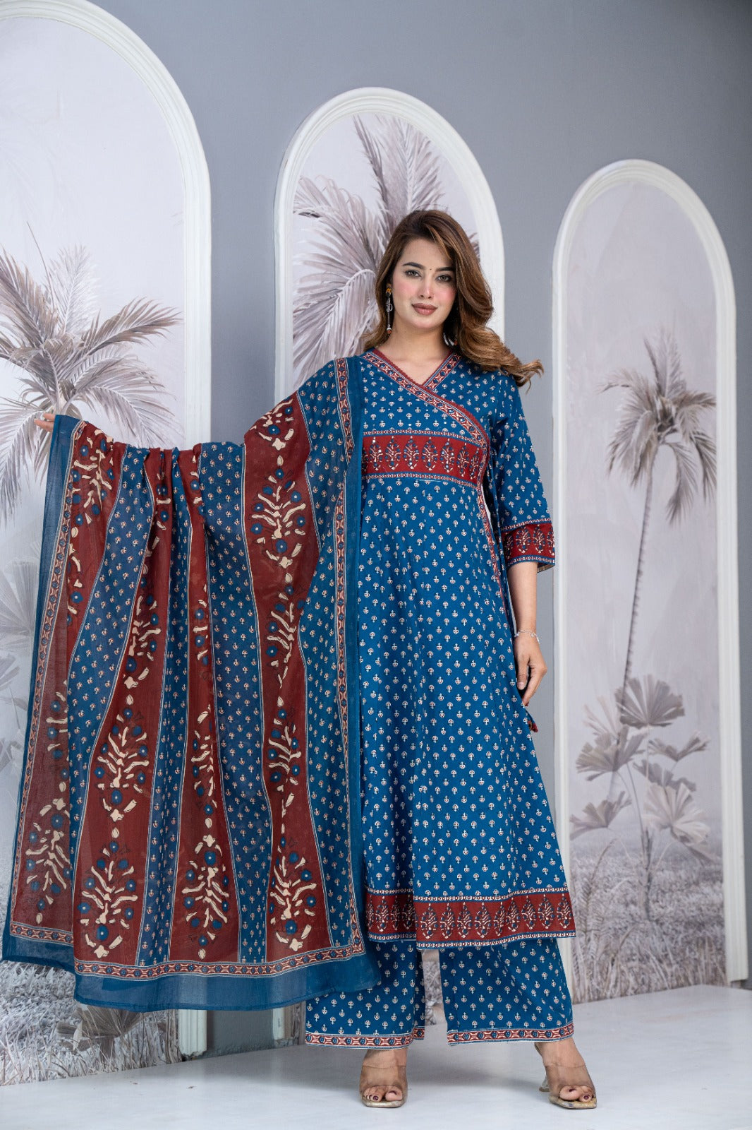 A woman models a traditional Indian salwar kameez set with a blue base, white polka dots, and a maroon border with floral prints, paired with a matching dupatta. She stands in front of a decorative backdrop with arched panels featuring palm tree illustrations.