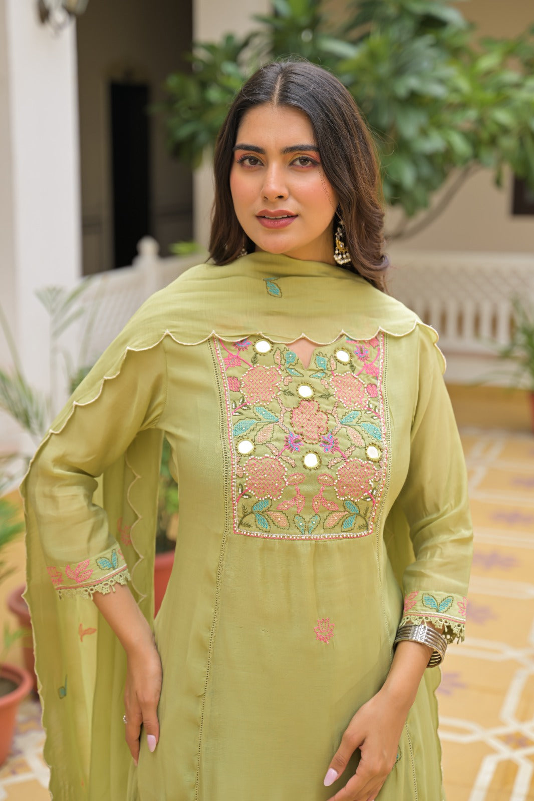 A woman wearing a light olive green traditional Indian salwar kameez suit with pink and green floral embroidery on the chest, standing in a courtyard setting.