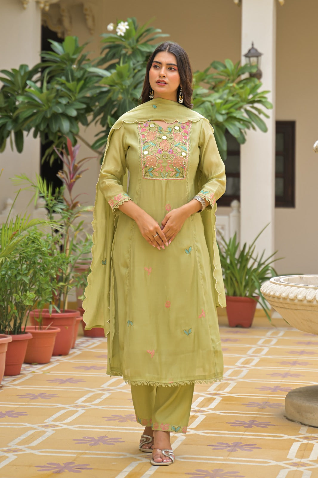 A woman stands in a courtyard wearing a light olive green traditional salwar kameez suit with colorful floral embroidery on the chest and scattered embroidered motifs on the kurta and dupatta.