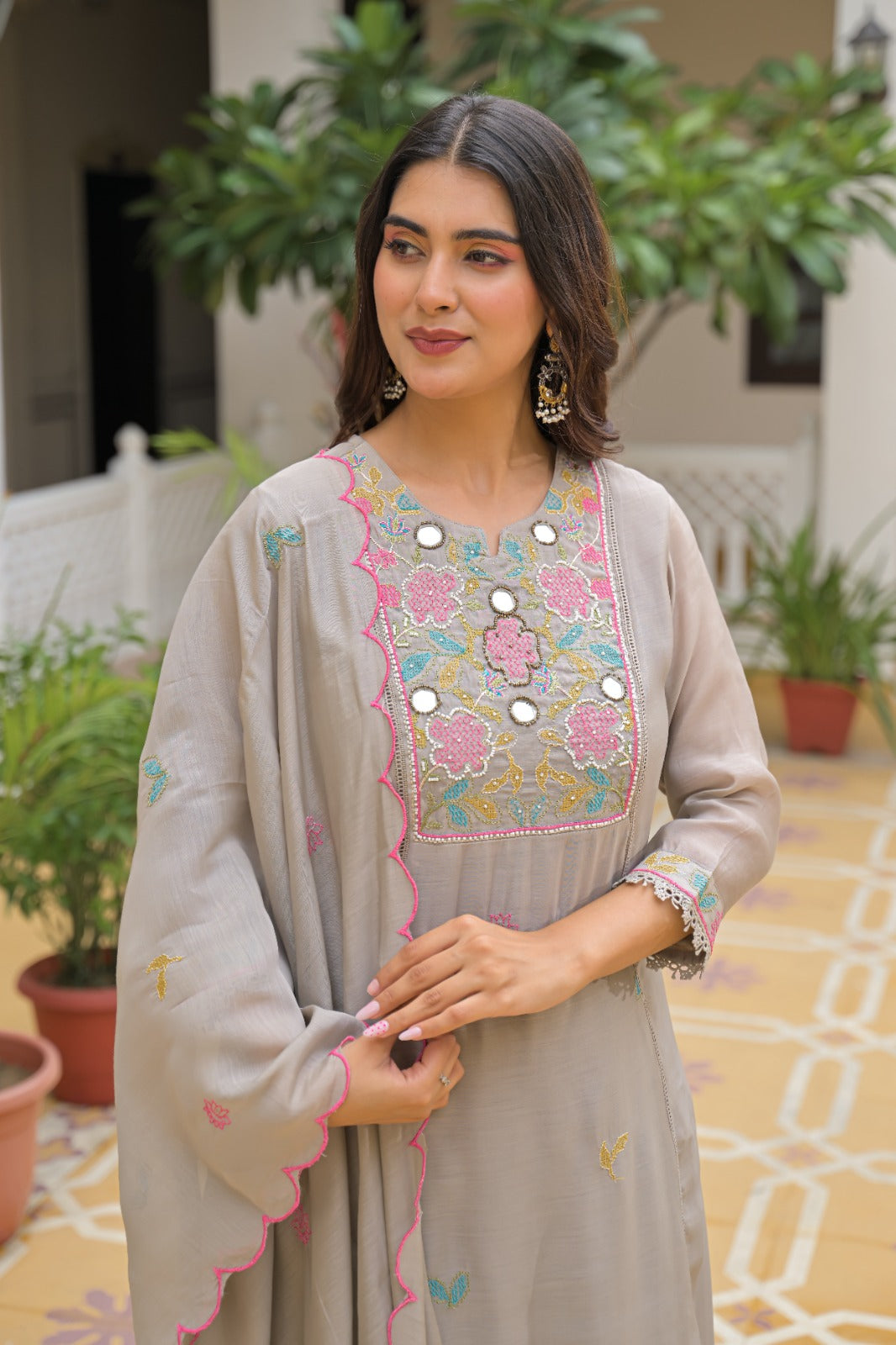 A woman wearing a light gray traditional embroidered kurta with pink, blue, and gold detailing, standing in a courtyard with potted plants.