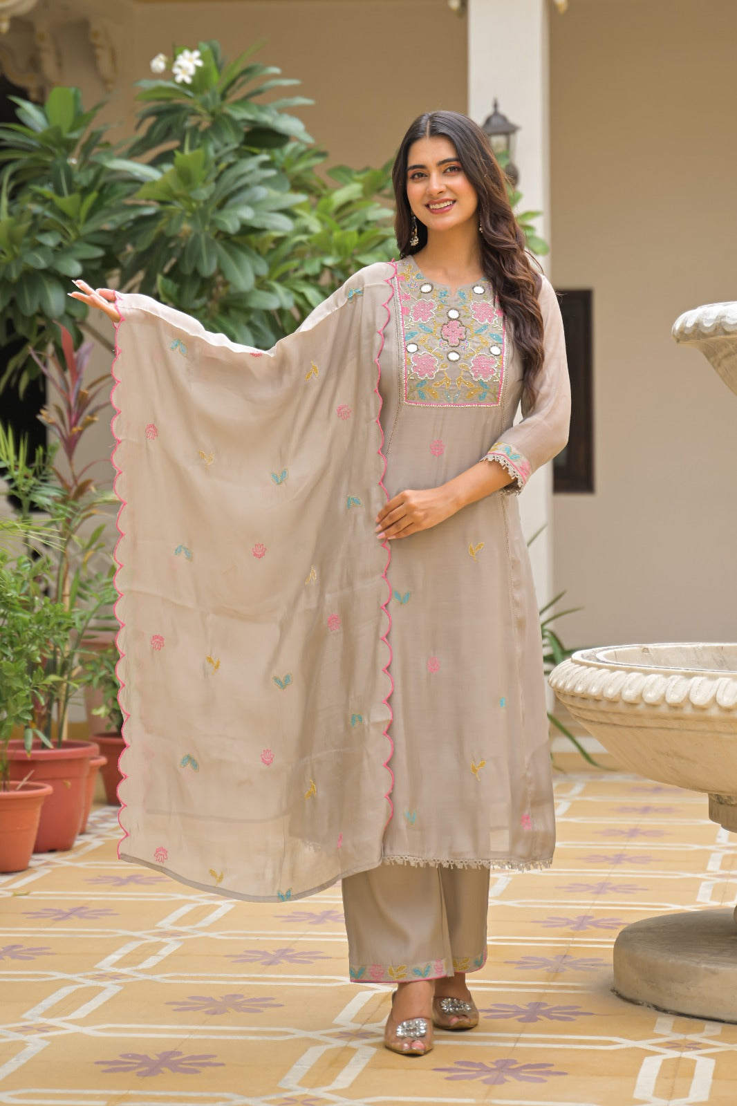 A woman models a traditional beige three-piece salwar kameez suit with a matching printed dupatta, standing in an outdoor courtyard setting with potted plants and a stone fountain.