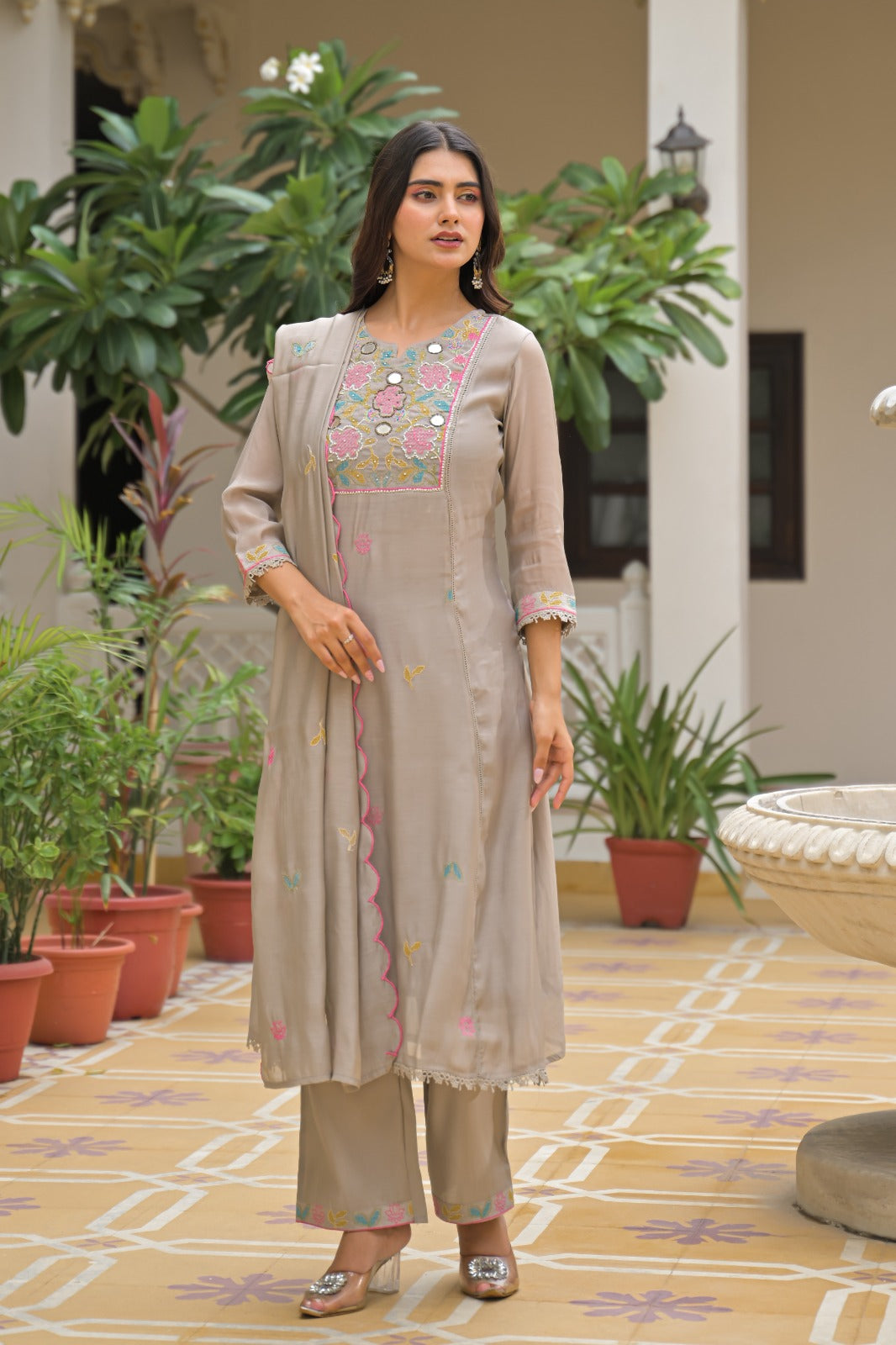 A woman models a traditional Indian salwar kameez suit set in a courtyard. The set consists of a light taupe-beige tunic with colorful floral embroidery on the chest and border, matching wide-leg trousers, and a coordinating embroidered jacket.