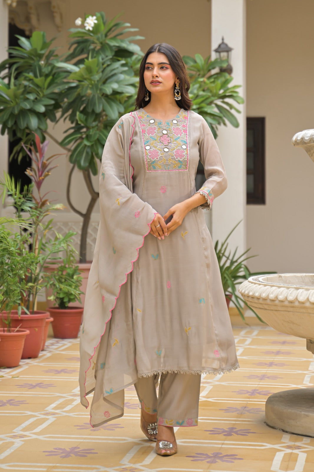 A woman models a light beige three-piece salwar kameez suit with colorful embroidery and a matching dupatta in an outdoor courtyard setting.