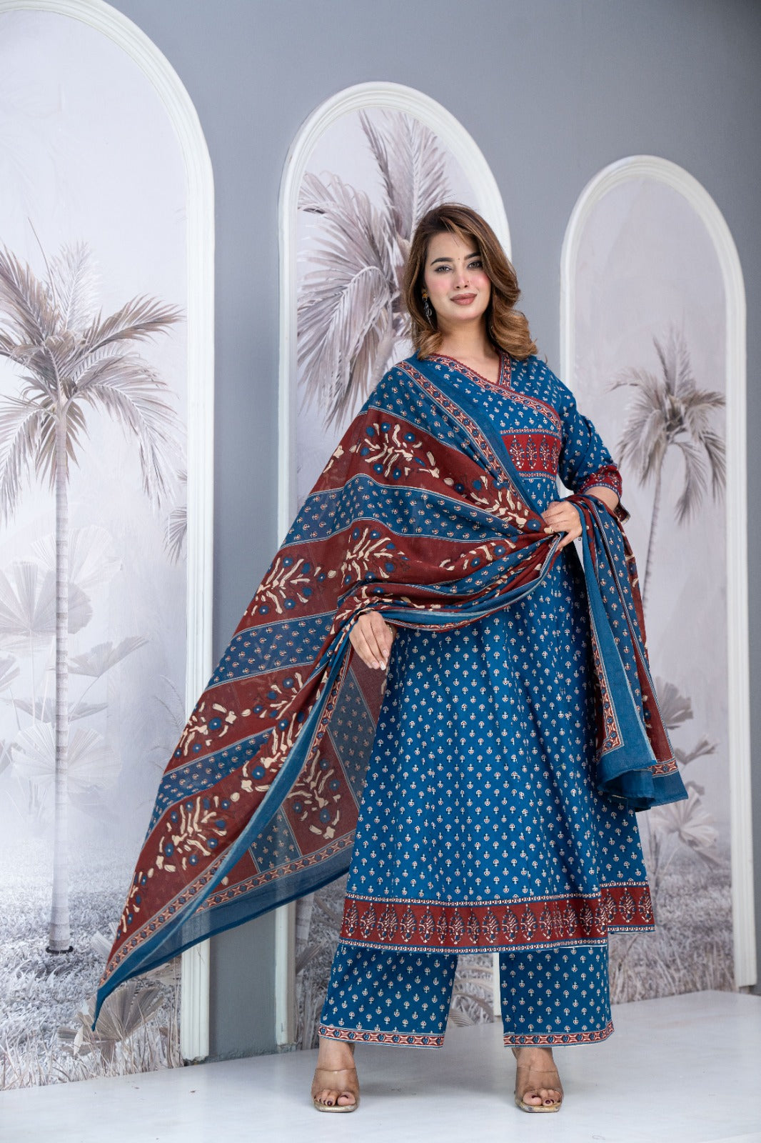 A woman models a traditional Indian salwar kameez set with a blue base, white polka dots, and a maroon border with floral prints, paired with a matching dupatta. She stands in front of a decorative backdrop with arched panels featuring palm tree illustrations.