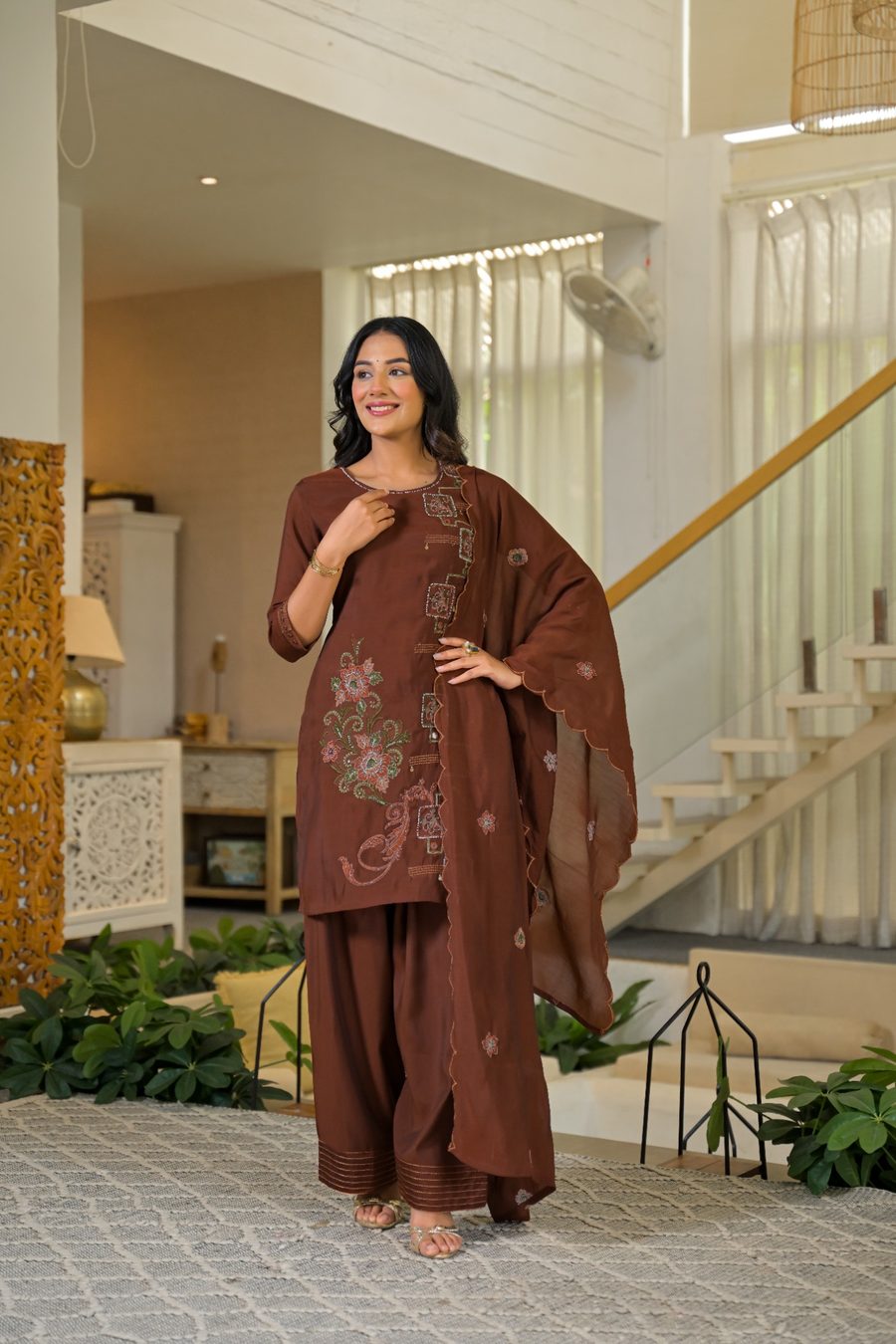 A woman modeling a brown three-piece salwar kameez suit with floral embroidery, standing in a modern, brightly lit indoor home setting with a staircase and plants in the background.