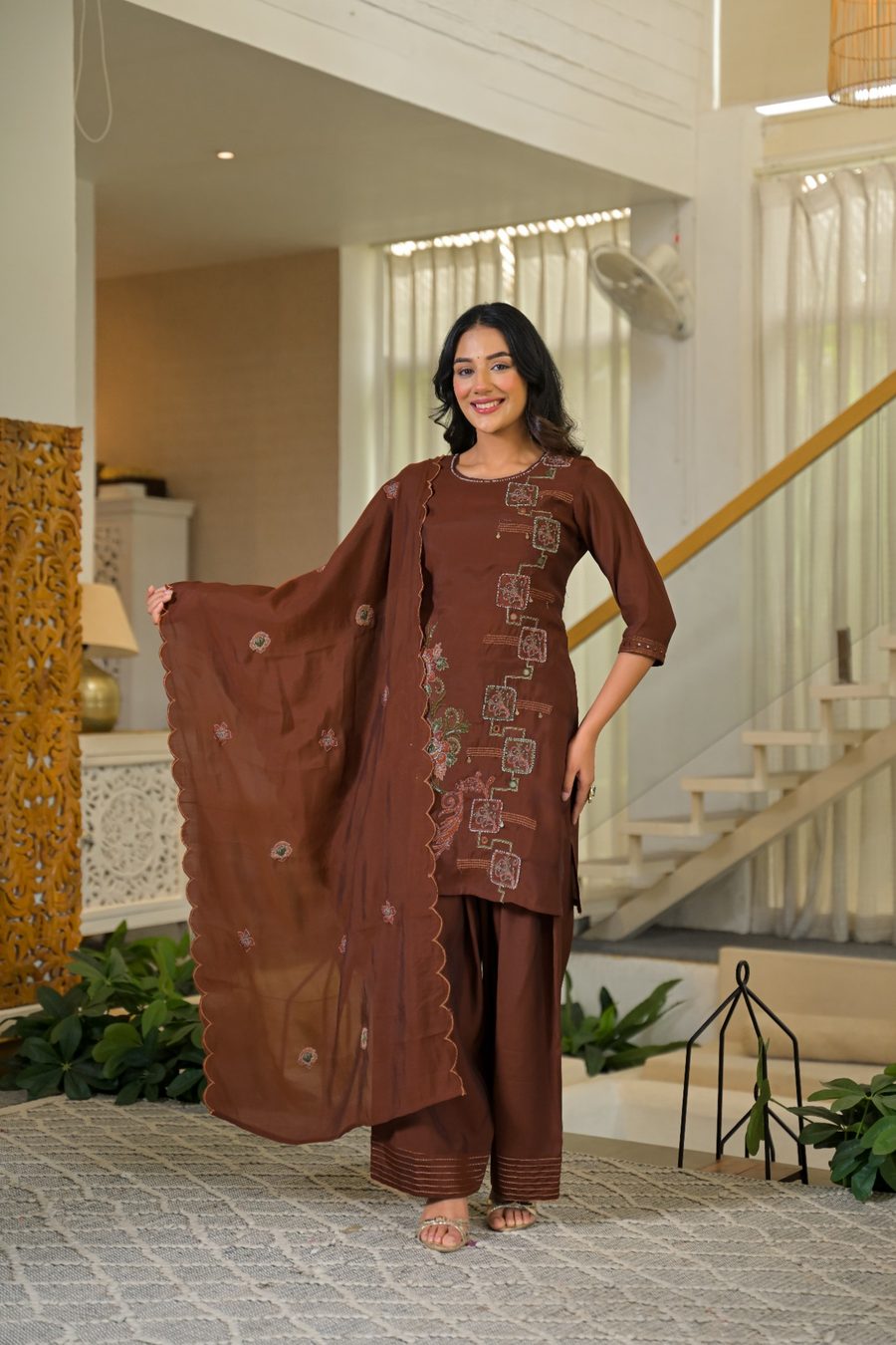 A woman modeling a brown three-piece salwar kameez suit with floral embroidery, standing in a modern, brightly lit indoor home setting with a staircase and plants in the background.